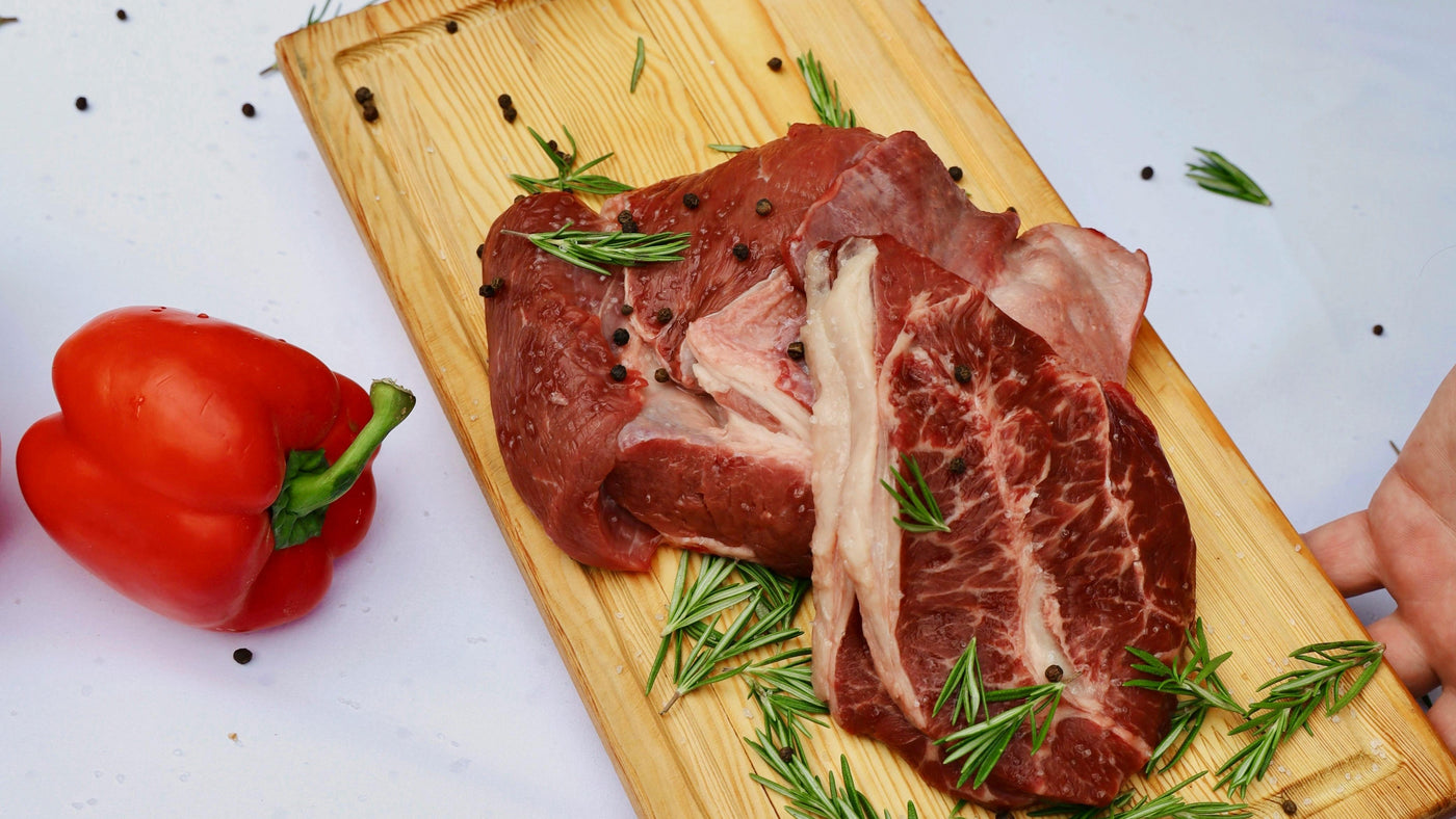 Raw meat on a wooden cutting board with rosemary, next to a red pepper on a white surface.
